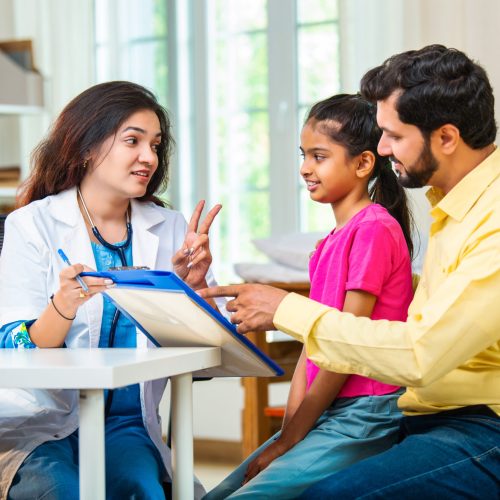Indian Asian woman doctor carefully writing a prescription for a cute little girl sitting with her father during a friendly medical checkup at a modern hospital or clinic, ensuring proper care