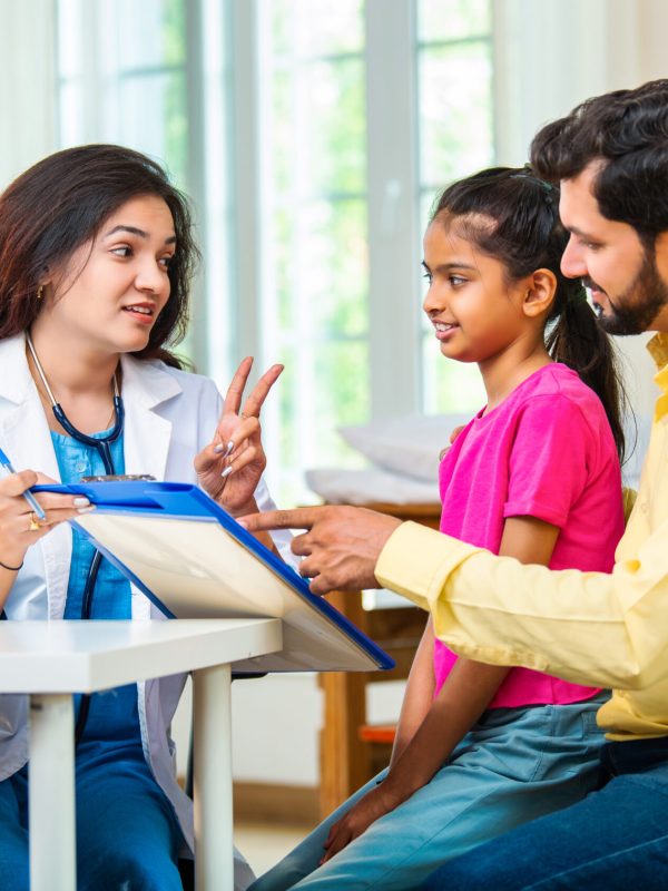 Indian Asian woman doctor carefully writing a prescription for a cute little girl sitting with her father during a friendly medical checkup at a modern hospital or clinic, ensuring proper care