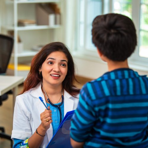 Indian Asian woman mental health counselor having a thoughtful conversation with a young boy patient sitting on a bed in modern clinic, assessing his general symptoms and providing emotional support