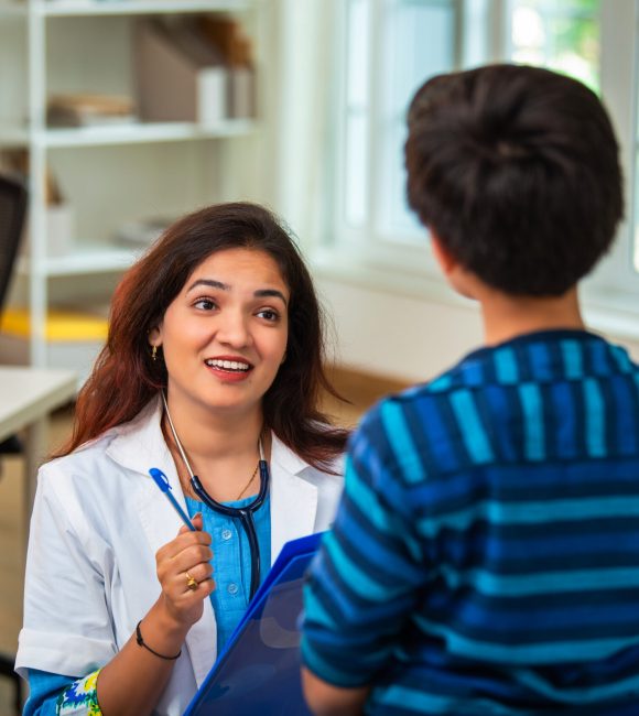 Indian Asian woman mental health counselor having a thoughtful conversation with a young boy patient sitting on a bed in modern clinic, assessing his general symptoms and providing emotional support